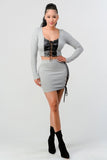 Grey two-piece set with cropped long sleeve top and mini skirt featuring black lace-up details on front and side. Model wearing black ankle boots.