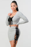 Grey two-piece set with cropped long sleeve shirt featuring lace-up PU leather detail and matching mini skirt with side lace-up accent, modeled by woman with dark hair in ponytail