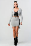 Grey two-piece set with cropped long sleeve shirt featuring black lace-up front detail and matching mini skirt with side lace-up accent, worn with black ankle boots